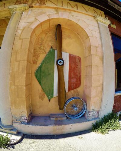 A close-up of an arched stone niche on a building facade displaying a wooden aircraft propeller, a shield painted with the Italian flag colors, and a spoked wheel, with the Latin inscription "ANIMVM" visible above.