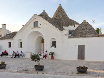 A large, white-washed trullo complex with multiple conical stone roofs and a prominent central archway, situated in a paved public square with people sitting on a bench and walking nearby.