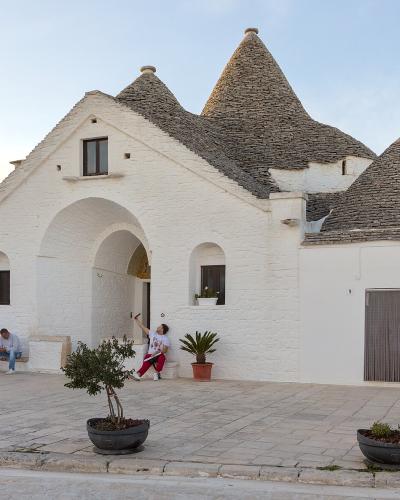 A large, white-washed trullo complex with multiple conical stone roofs and a prominent central archway, situated in a paved public square with people sitting on a bench and walking nearby.
