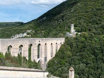 A long, high medieval stone aqueduct bridge with multiple tall arches and piers, set against a densely forested hillside under a partly cloudy sky; a small tower-like structure is visible on the hill to the right, and part of a foreground wall or