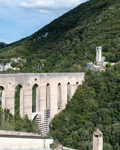 A long, high medieval stone aqueduct bridge with multiple tall arches and piers, set against a densely forested hillside under a partly cloudy sky; a small tower-like structure is visible on the hill to the right, and part of a foreground wall or