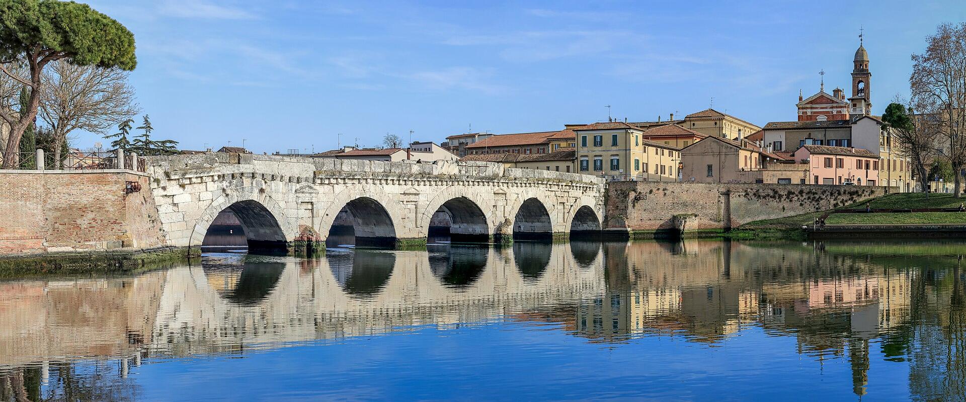 A panoramic photograph of the ancient Roman stone arch bridge known as the Ponte di Tiberio, spanning a calm river with clear reflections, set against a backdrop of historic Italian buildings and a church tower under a blue sky.