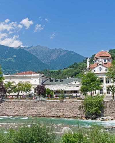 A wide-angle view of the Kurhaus Merano complex, featuring its distinctive rotunda and white facade, situated along the bank of a rushing river with mountains in the background.