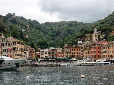 A panoramic view of a crescent-shaped harbor lined with colorful pastel buildings, luxury yachts moored in the foreground, and a hillside covered in greenery and villas, including a prominent church tower.