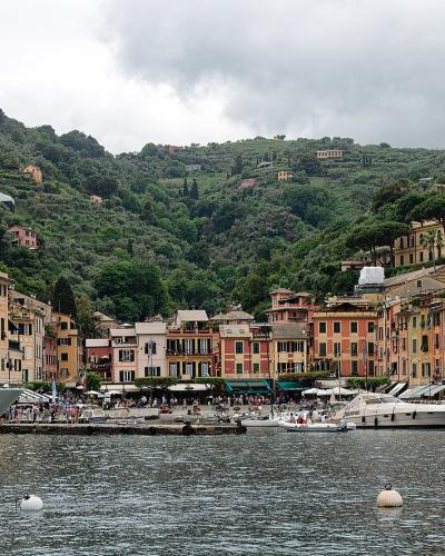A panoramic view of a crescent-shaped harbor lined with colorful pastel buildings, luxury yachts moored in the foreground, and a hillside covered in greenery and villas, including a prominent church tower.
