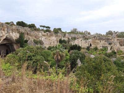 A wide landscape view of a large, excavated limestone quarry with steep rock faces, numerous caves and openings, and dense Mediterranean vegetation in the foreground and along the rim.