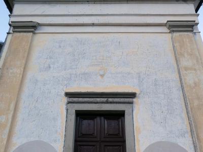 The image shows the facade of a small, weathered church with a triangular pediment, a central dark wooden door, and a circular oculus window near the roofline, set against a clear sky.