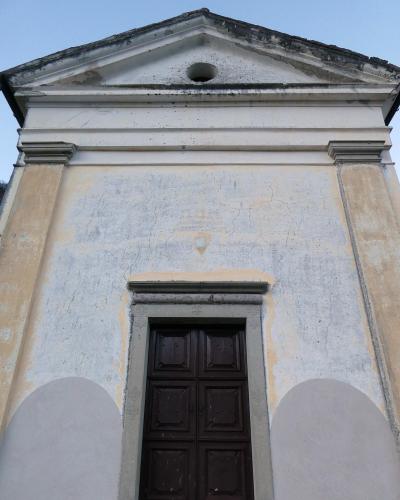 The image shows the facade of a small, weathered church with a triangular pediment, a central dark wooden door, and a circular oculus window near the roofline, set against a clear sky.