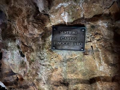 A close-up of a dark metal plaque mounted on a rough, textured limestone cave wall, inscribed with the text "SENTIERO CARLO FINOCCHIARO".