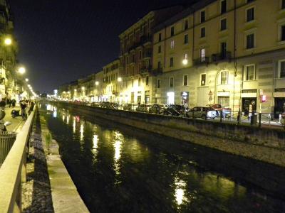 A night-time photograph of a straight, urban canal lined with multi-story buildings, streetlights, parked cars, and a pedestrian walkway on the left.