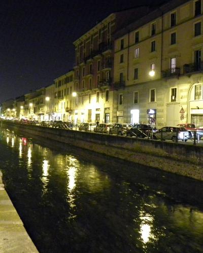 A night-time photograph of a straight, urban canal lined with multi-story buildings, streetlights, parked cars, and a pedestrian walkway on the left.