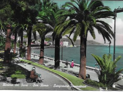 A vintage colorized postcard showing a curved seaside promenade lined with tall palm trees, a stone balustrade overlooking the ocean, and people strolling or sitting on benches, with a town visible on the hillside in the background.