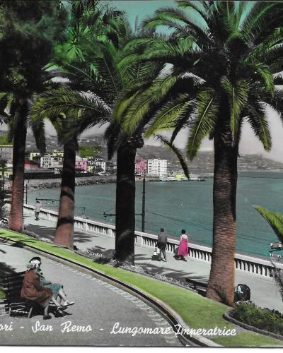 A vintage colorized postcard showing a curved seaside promenade lined with tall palm trees, a stone balustrade overlooking the ocean, and people strolling or sitting on benches, with a town visible on the hillside in the background.