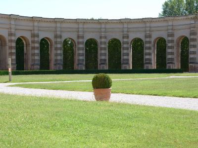 A long, curved, two-story loggia with rusticated pilasters and arched openings, fronting a manicured lawn with gravel paths and potted topiary.