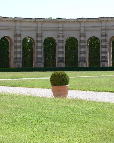 A long, curved, two-story loggia with rusticated pilasters and arched openings, fronting a manicured lawn with gravel paths and potted topiary.