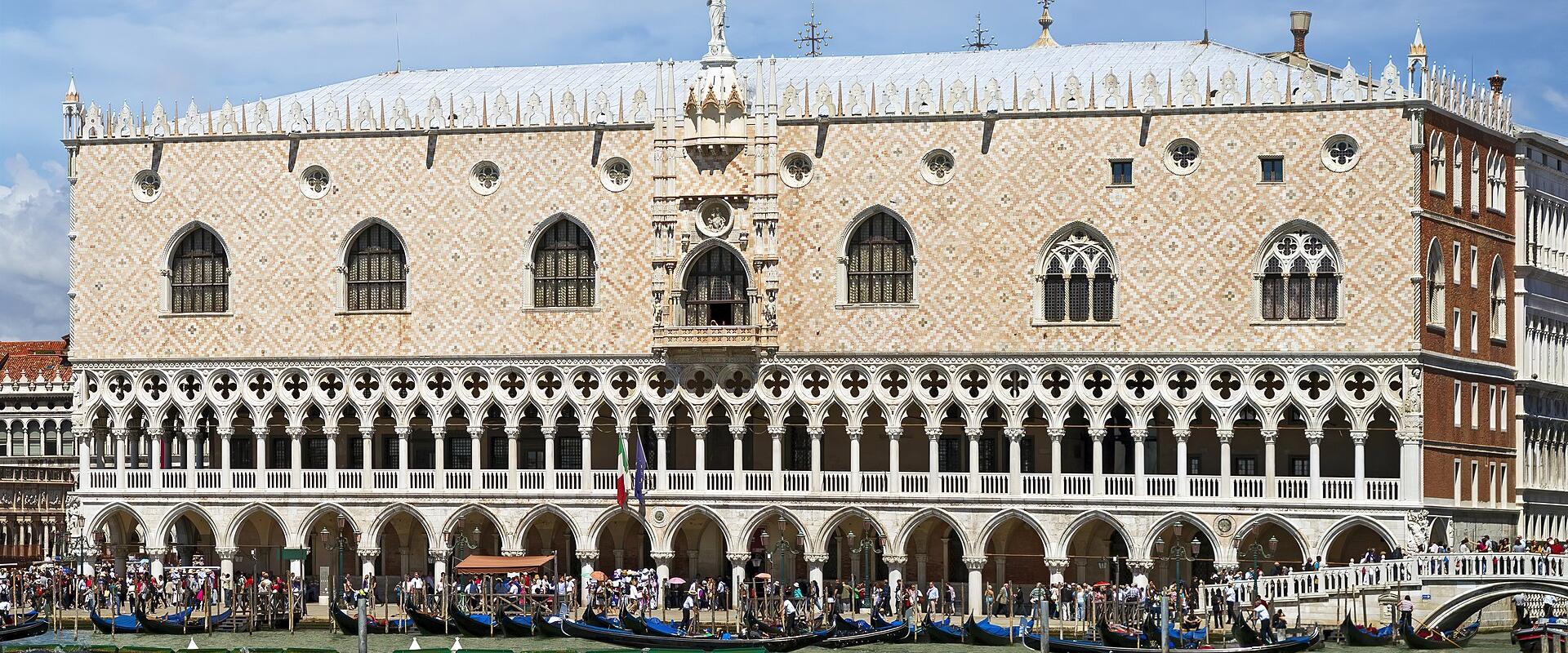 The image shows the Doge’s Palace in Venice, Italy, viewed from the water with gondolas moored along the waterfront and tourists visible on the promenade; its distinctive Gothic architecture with diamond-patterned facade, arched loggias, and ornat