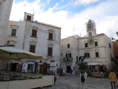 A sunlit European-style town square with white-washed buildings, outdoor café seating under umbrellas, a prominent clock tower on the right building, and people walking or sitting in the paved plaza.