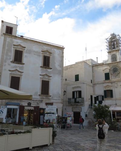 A sunlit European-style town square with white-washed buildings, outdoor café seating under umbrellas, a prominent clock tower on the right building, and people walking or sitting in the paved plaza.