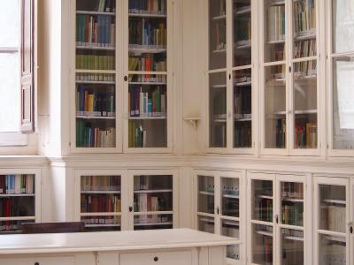 An interior room with white built-in bookshelves filled with books, a large white table, and wooden chairs, likely a library or reading room.
