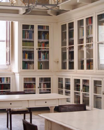 An interior room with white built-in bookshelves filled with books, a large white table, and wooden chairs, likely a library or reading room.