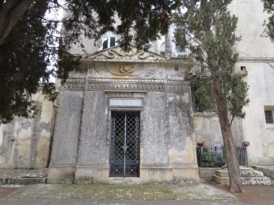 The image shows a weathered, ornate stone mausoleum or tomb structure with a gated entrance, set within a cemetery environment with trees and adjacent buildings.