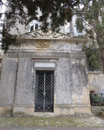 The image shows a weathered, ornate stone mausoleum or tomb structure with a gated entrance, set within a cemetery environment with trees and adjacent buildings.