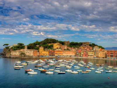 A panoramic view of a crescent-shaped bay filled with numerous small boats, backed by a dense cluster of colorful buildings and a hilltop fortress under a cloudy sky.