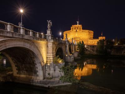 A night photograph showing the illuminated cylindrical fortress of Castel Sant'Angelo in the background, connected to the foreground by the Ponte Sant'Angelo bridge, which is lined with angel statues, all reflected in the Tiber River.
