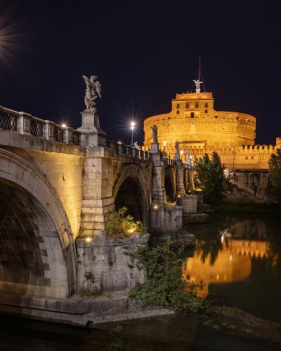 A night photograph showing the illuminated cylindrical fortress of Castel Sant'Angelo in the background, connected to the foreground by the Ponte Sant'Angelo bridge, which is lined with angel statues, all reflected in the Tiber River.