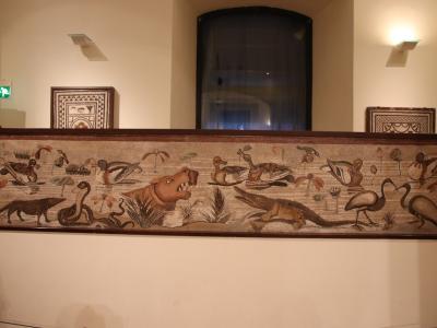 A long, rectangular mosaic panel depicting a riverine scene with animals including a hippopotamus, crocodile, snake, dog, ducks, and ibises, displayed horizontally on a white plinth in a museum gallery with other framed mosaic fragments on the wal