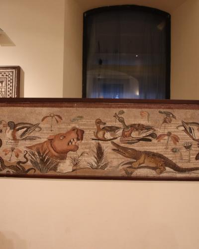 A long, rectangular mosaic panel depicting a riverine scene with animals including a hippopotamus, crocodile, snake, dog, ducks, and ibises, displayed horizontally on a white plinth in a museum gallery with other framed mosaic fragments on the wal