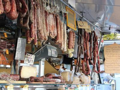A market stall displaying hanging cured sausages and salami, with handwritten Italian signs for products like "Peperino," "Fegato," and "Zafferano," along with a digital scale and cheese display.