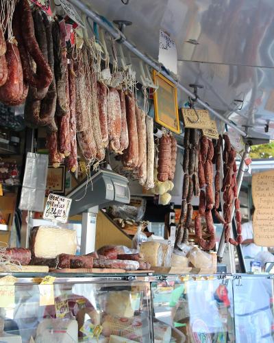 A market stall displaying hanging cured sausages and salami, with handwritten Italian signs for products like "Peperino," "Fegato," and "Zafferano," along with a digital scale and cheese display.