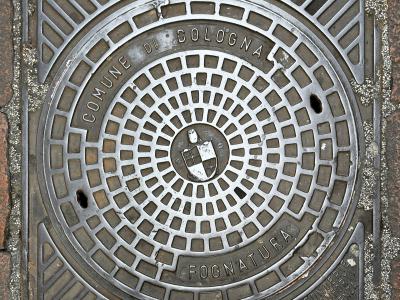 A close-up of a square metal manhole cover set into a paved street, featuring the text "COMUNE DI BOLOGNA" and "FOGNATURA" (sewer), along with the city's coat of arms in the center.
