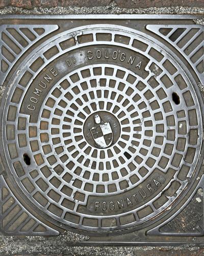 A close-up of a square metal manhole cover set into a paved street, featuring the text "COMUNE DI BOLOGNA" and "FOGNATURA" (sewer), along with the city's coat of arms in the center.
