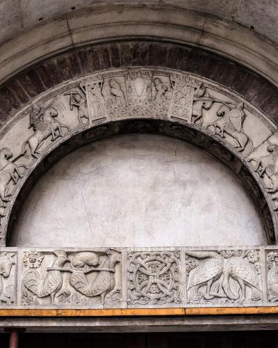 A close-up photograph of a Romanesque stone portal featuring an archivolt with sculpted knights on horseback and a lintel with various animal and mythological carvings.