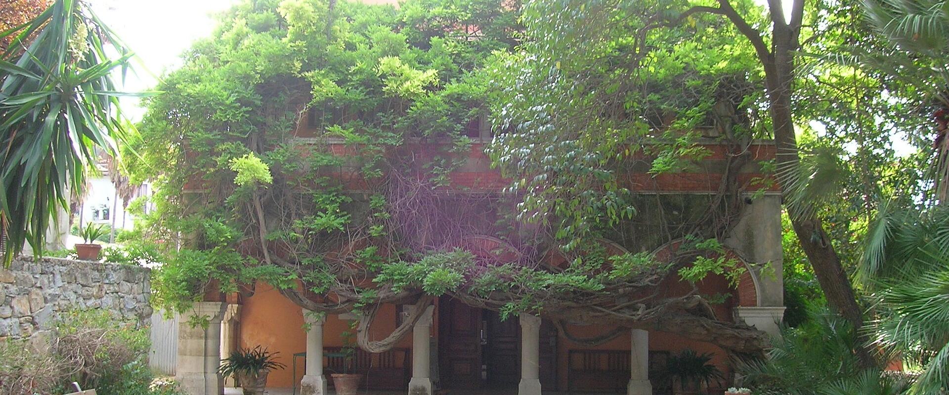 A two-story terracotta-colored building with a covered loggia supported by stone columns, heavily overgrown with green vines and trees, set within a gravel courtyard with wooden benches and potted plants.