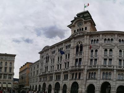 The image shows the Palazzo del Governo (Government Palace) in Trieste, Italy, a grand neoclassical building with a clock tower and Italian flag, situated at Piazza Unità d’Italia, with crowds and adjacent buildings visible under an overcast sky.