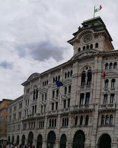 The image shows the Palazzo del Governo (Government Palace) in Trieste, Italy, a grand neoclassical building with a clock tower and Italian flag, situated at Piazza Unità d’Italia, with crowds and adjacent buildings visible under an overcast sky.