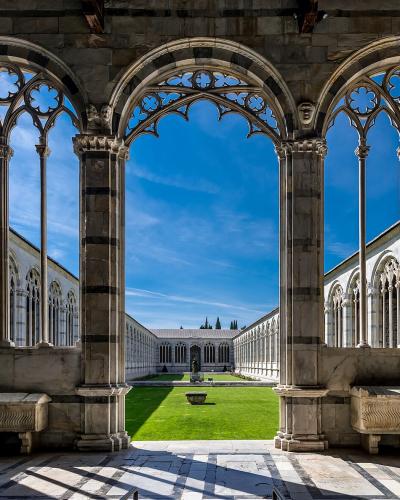 The image shows the interior of a Gothic-style cloister with pointed arches, slender columns, and decorative tracery, looking out onto a grassy courtyard with stone sarcophagi; the architecture features black-and-white striped marble typical of Tu