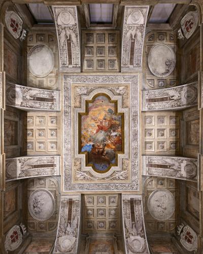 A high-angle photograph of an ornate, coffered ceiling featuring a central fresco surrounded by trompe-l'œil architectural elements, pilasters with eagles, and red-and-white heraldic shields.