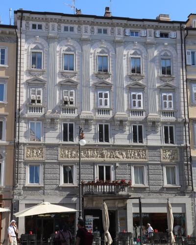 A multi-story, light-colored neoclassical building with ornate window surrounds, pilasters, and a sculpted frieze above the ground floor; storefronts at street level include “COVERGOO” and “golden point”; people are walking or sitting at outdoor c