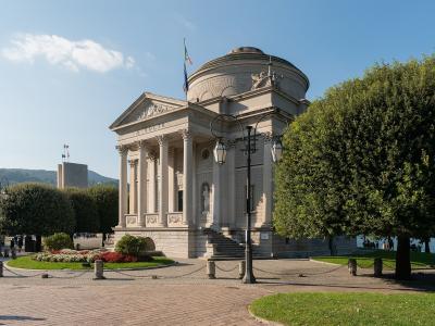 A neoclassical building with a portico of columns, a pediment inscribed "A. VOLTA", a domed rotunda, and an Italian flag on top, situated in a landscaped plaza near water with trees and lampposts.