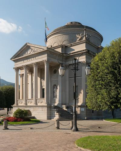 A neoclassical building with a portico of columns, a pediment inscribed "A. VOLTA", a domed rotunda, and an Italian flag on top, situated in a landscaped plaza near water with trees and lampposts.