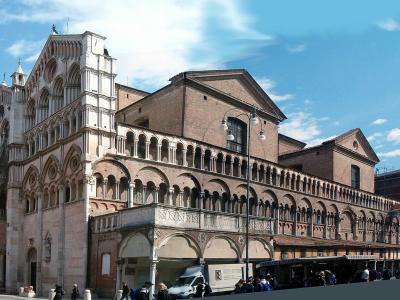 A wide-angle photograph of a large, ornate cathedral with a distinctive pink-and-white striped facade, a long brick side arcade, and a separate bell tower under scaffolding, set in a public square with people and vehicles.