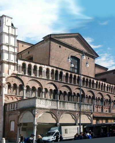 A wide-angle photograph of a large, ornate cathedral with a distinctive pink-and-white striped facade, a long brick side arcade, and a separate bell tower under scaffolding, set in a public square with people and vehicles.