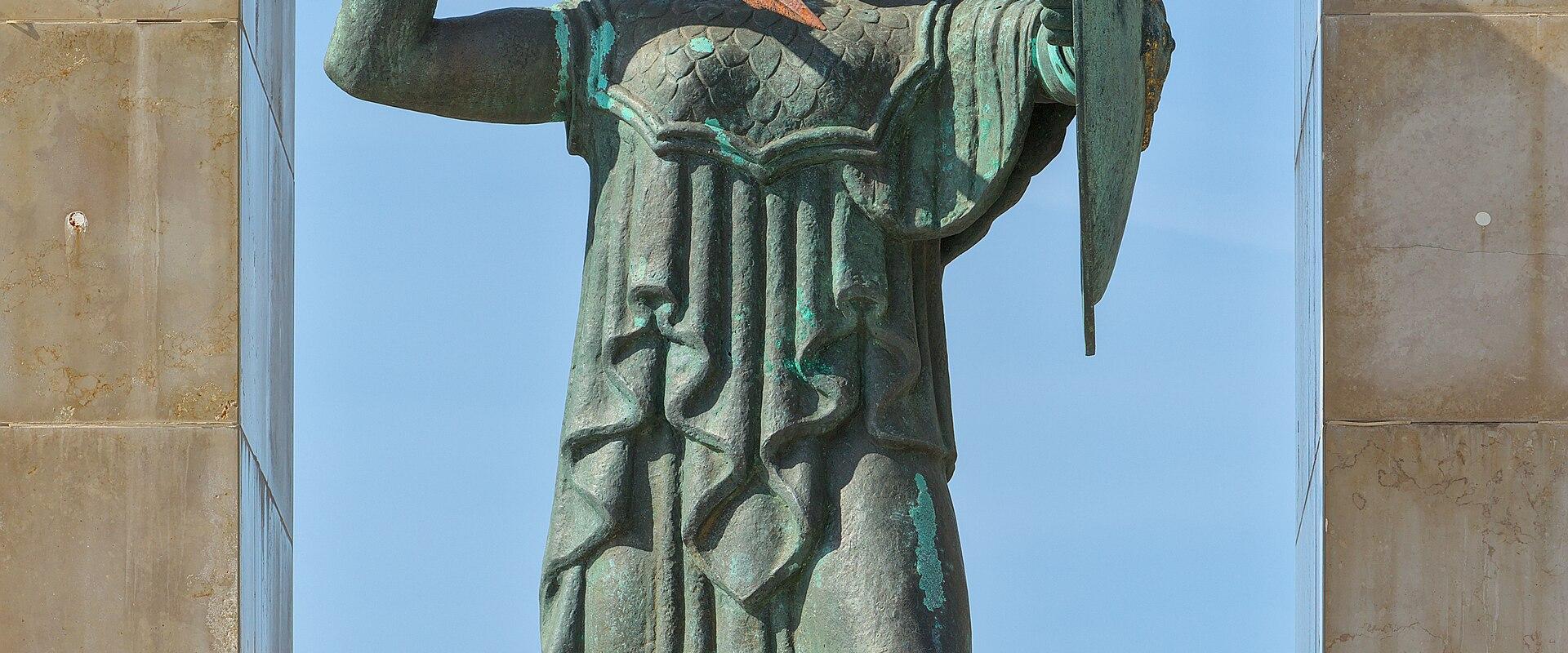 A bronze statue of a female warrior holding a spear and shield, standing on a pedestal between two tall stone pillars under a clear blue sky.