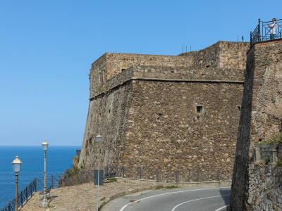 A stone fortress wall overlooking the sea, with a paved walkway, lampposts, and a person on an upper terrace.