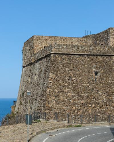 A stone fortress wall overlooking the sea, with a paved walkway, lampposts, and a person on an upper terrace.
