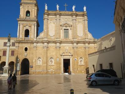 A large, light-colored stone cathedral with a single bell tower on the left, a central facade featuring statues along the roofline and niches with statues below, situated in a paved public square under a clear blue sky.